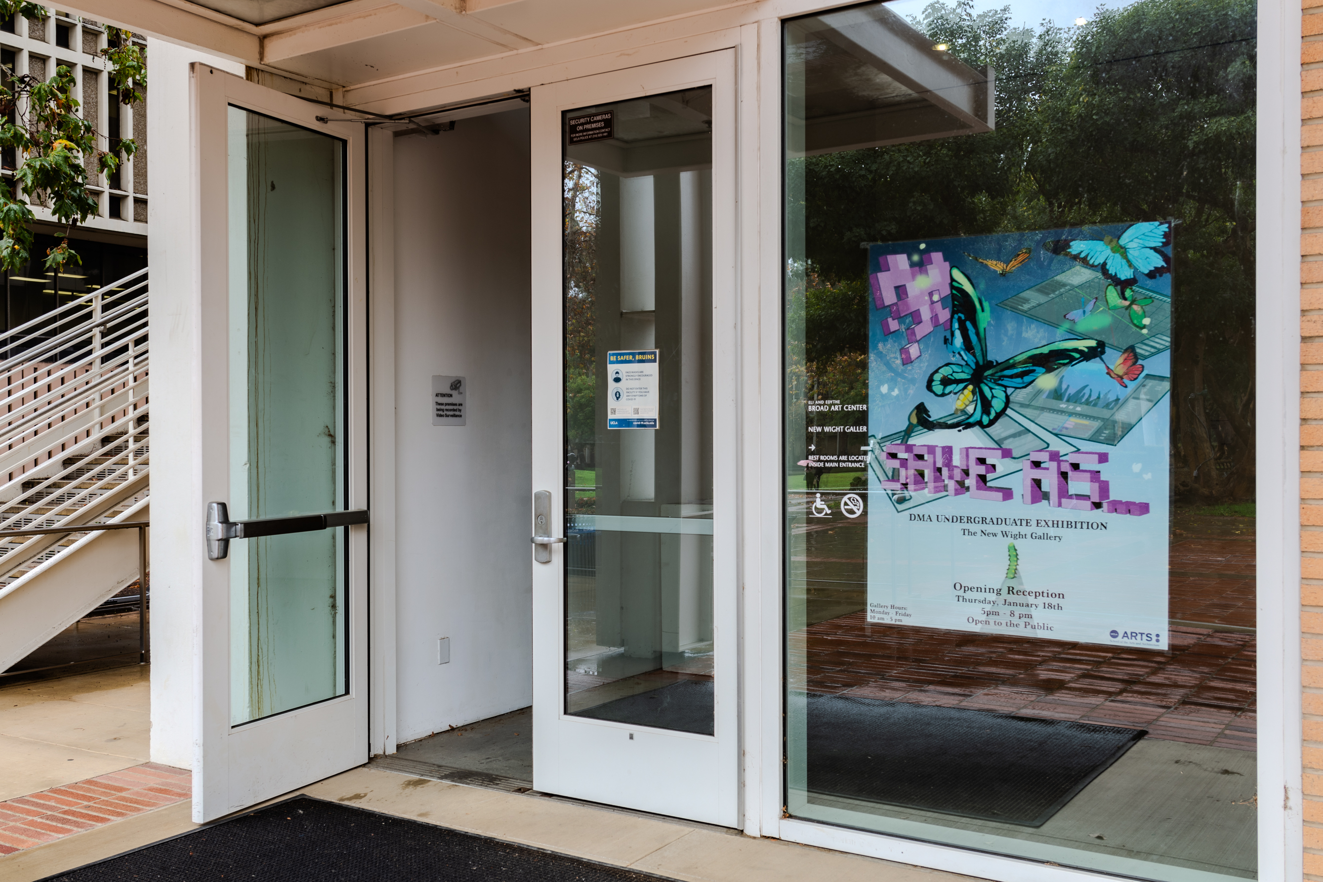 Exterior entrance to a gallery with a printed poster hung on a glass window to the left. On the right is two white doors with one opened towards the gallery.